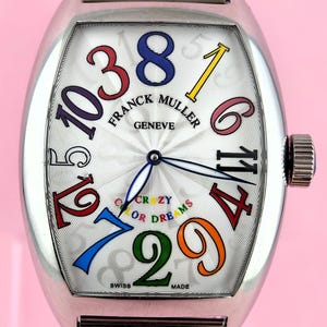 Op de afbeelding: Een zilveren Franck Muller Crazy Hours horloge met een witte wijzerplaat en kleurrijke cijfers. Het horloge heeft een uniek ontwerp met de woorden "CRAZY OR DREAMS" en "SWISS MADE" op de wijzerplaat. Het horloge heeft een zilveren band en een kroon.