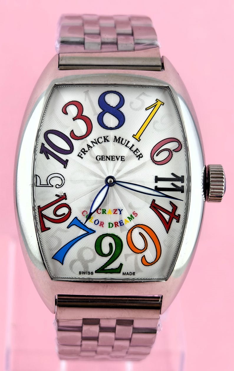 May include: A silver Franck Muller Crazy Hours watch with a white face and colorful numerals. The watch features a unique design with the words "CRAZY OR DREAMS" and "SWISS MADE" on the face. The watch has a silver band and a crown.