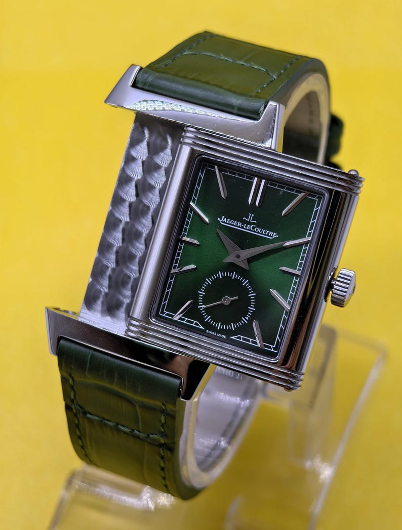 May include: A rectangular silver-tone wristwatch with a green sunray dial, silver hands, and indices. The watch features a small seconds sub-dial, the text Jaeger-LeCoultre, and a green alligator-textured leather strap with a metallic side panel.