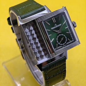 May include: A rectangular silver-tone wristwatch featuring a sunburst green dial with the Jaeger-LeCoultre logo and text, a small seconds sub-dial, and a textured metallic side panel. The watch is attached to a dark green alligator-patterned leather strap.