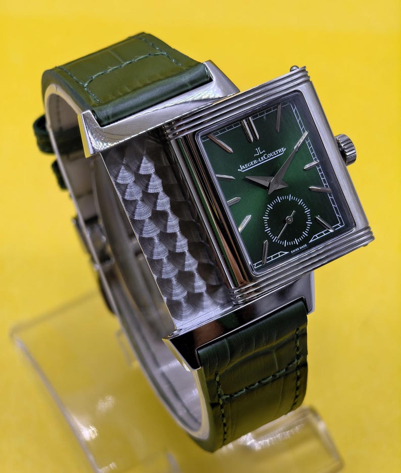 May include: A rectangular silver-tone wristwatch featuring a sunburst green dial with the Jaeger-LeCoultre logo and text, a small seconds sub-dial, and a textured metallic side panel. The watch is attached to a dark green alligator-patterned leather strap.