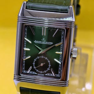 May include: A rectangular silver-tone wristwatch with a dark green dial, silver hands, and silver hour markers. The watch features the text Jaeger-LeCoultre and Swiss Made on the face, with a small sub-dial and a dark green alligator-textured leather strap.