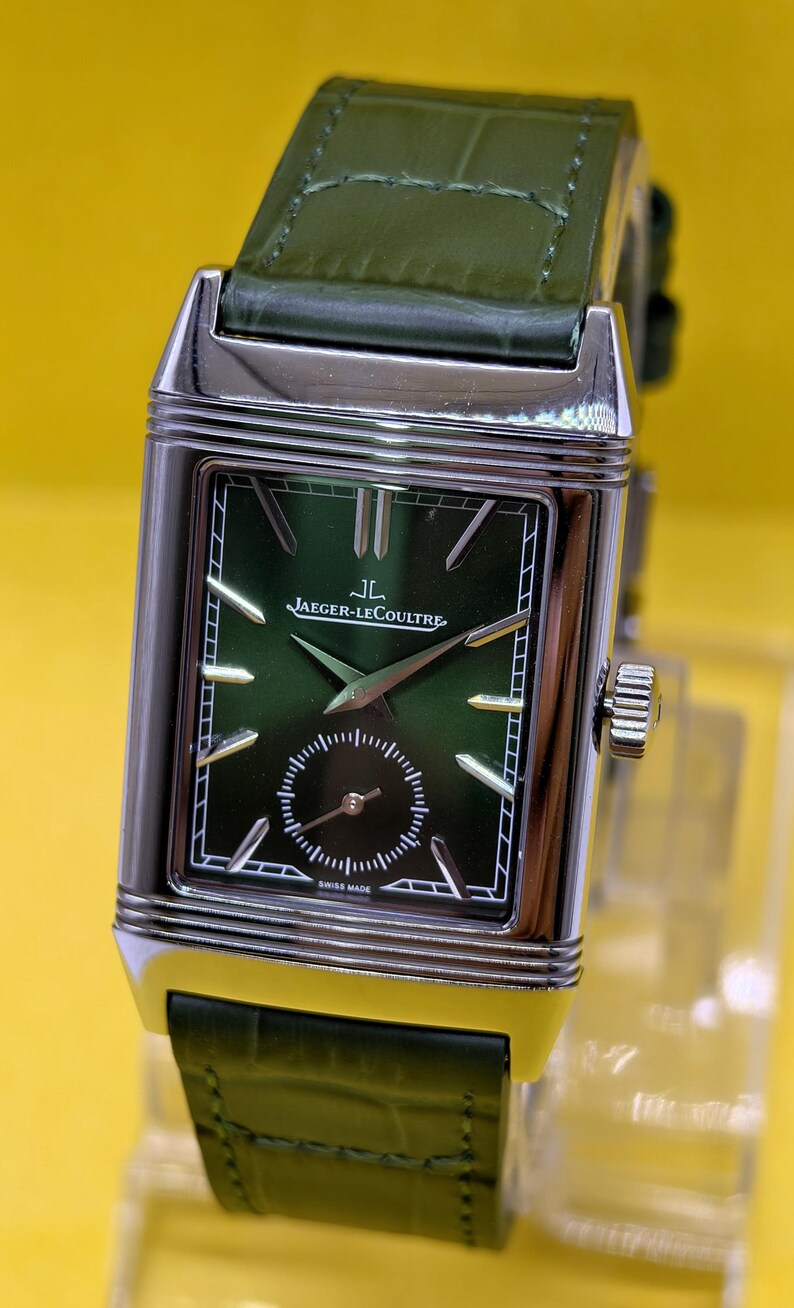 May include: A rectangular silver-tone wristwatch with a dark green dial, silver hands, and silver hour markers. The watch features the text Jaeger-LeCoultre and Swiss Made on the face, with a small sub-dial and a dark green alligator-textured leather strap.