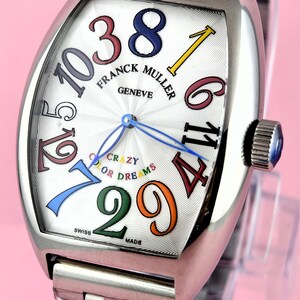 May include: A stainless steel Franck Muller Geneve watch with a white face and colorful numerals. The watch features the text "CRAZY OR DREAMS" and a stainless steel band. The watch is Swiss made.