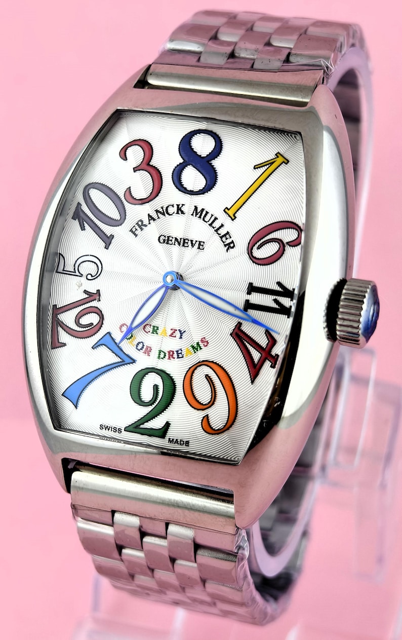 May include: A stainless steel Franck Muller Geneve watch with a white face and colorful numerals. The watch features the text "CRAZY OR DREAMS" and a stainless steel band. The watch is Swiss made.