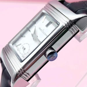 Pu&ograve; includere: Un orologio da polso rettangolare argentato con quadrante bianco e cinturino in pelle nera. L'orologio presenta un piccolo quadrante dei secondi e lancette argentate. La corona &egrave; blu. L'orologio &egrave; angolato, mostrando il suo design.
