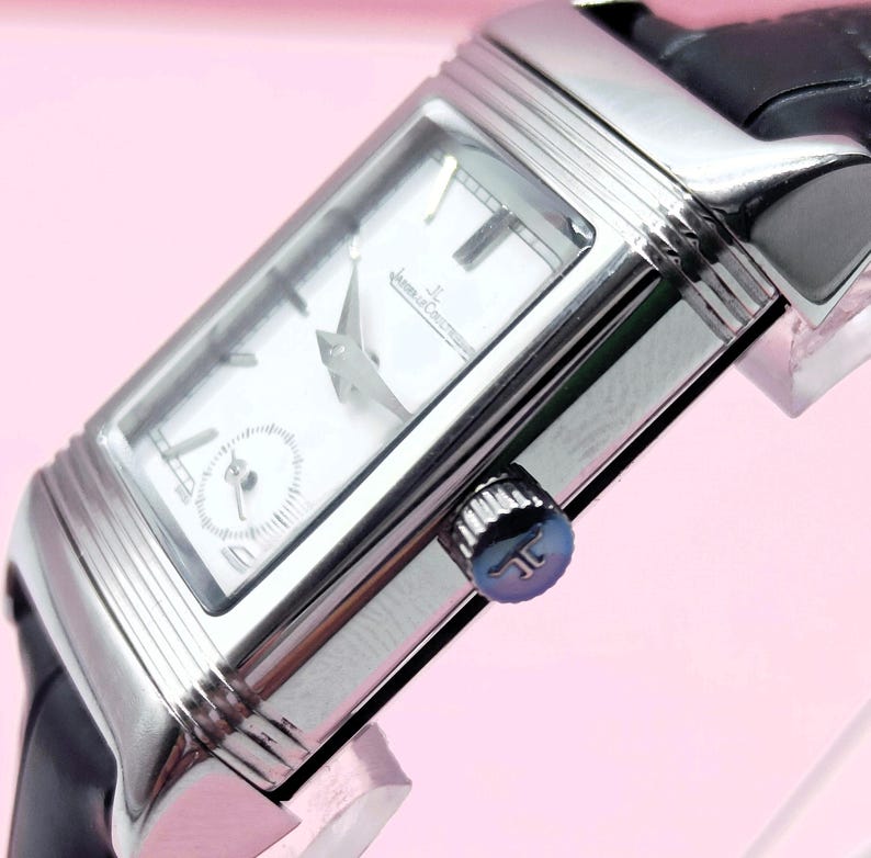 Pu&ograve; includere: Un orologio da polso rettangolare argentato con quadrante bianco e cinturino in pelle nera. L'orologio presenta un piccolo quadrante dei secondi e lancette argentate. La corona &egrave; blu. L'orologio &egrave; angolato, mostrando il suo design.