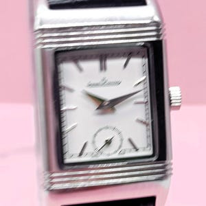 Pu&ograve; includere: Un orologio da polso rettangolare argentato con quadrante bianco e cinturino in pelle nera. Il quadrante dell'orologio presenta lancette nere e un piccolo quadrante dei secondi. Il marchio "Jaeger-LeCoultre" &egrave; stampato sul quadrante.