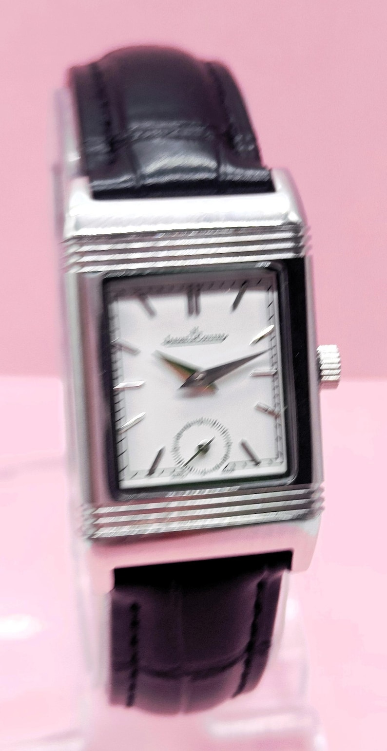 Pu&ograve; includere: Un orologio da polso rettangolare argentato con quadrante bianco e cinturino in pelle nera. Il quadrante dell'orologio presenta lancette nere e un piccolo quadrante dei secondi. Il marchio "Jaeger-LeCoultre" &egrave; stampato sul quadrante.