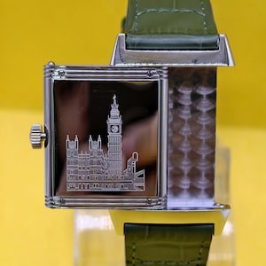 May include: A rectangular silver-tone wristwatch with a reversible case featuring an engraved illustration of the Big Ben clock tower and the Houses of Parliament on the polished metal surface, paired with a textured olive green leather strap.