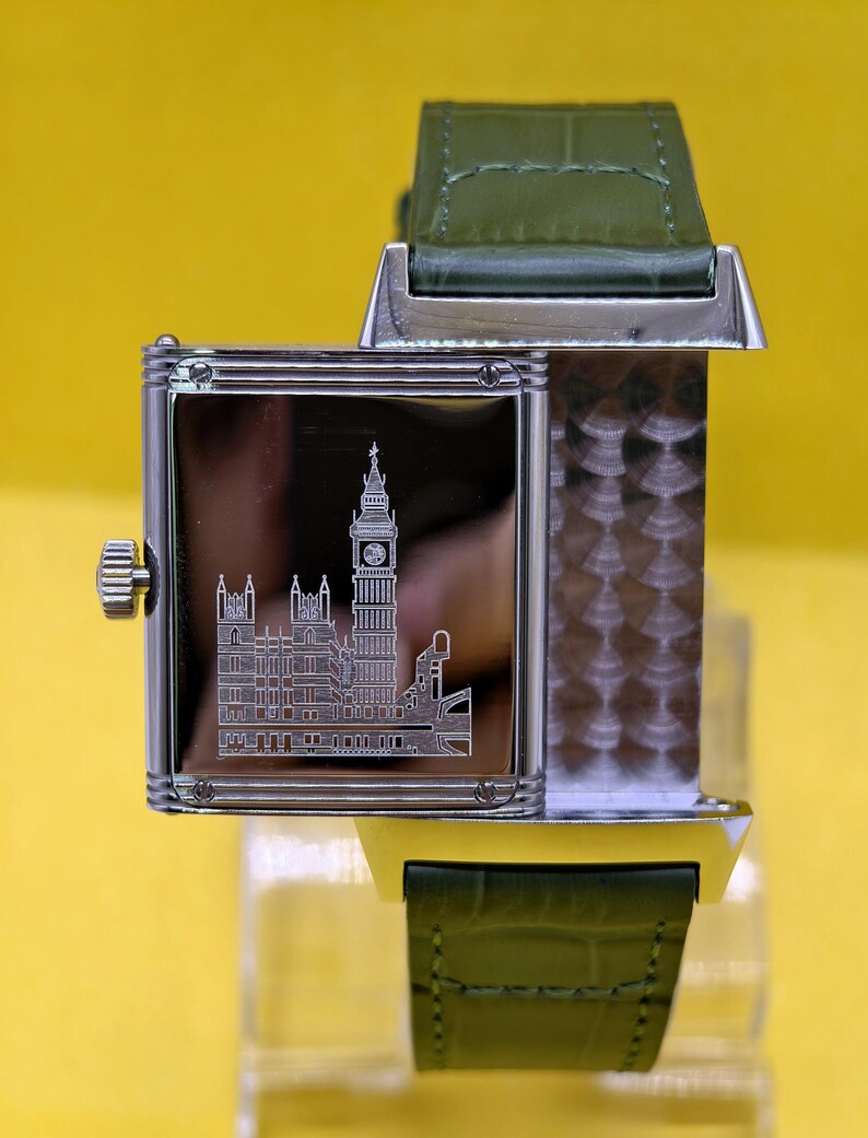 May include: A rectangular silver-tone wristwatch with a reversible case featuring an engraved illustration of the Big Ben clock tower and the Houses of Parliament on the polished metal surface, paired with a textured olive green leather strap.
