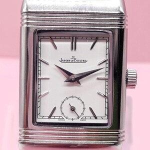Pu&ograve; includere: Un orologio Jaeger-LeCoultre rettangolare argentato con quadrante bianco e lancette nere. L'orologio presenta un piccolo quadrante dei secondi e un cinturino in pelle nera. Il quadrante mostra il logo Jaeger-LeCoultre.
