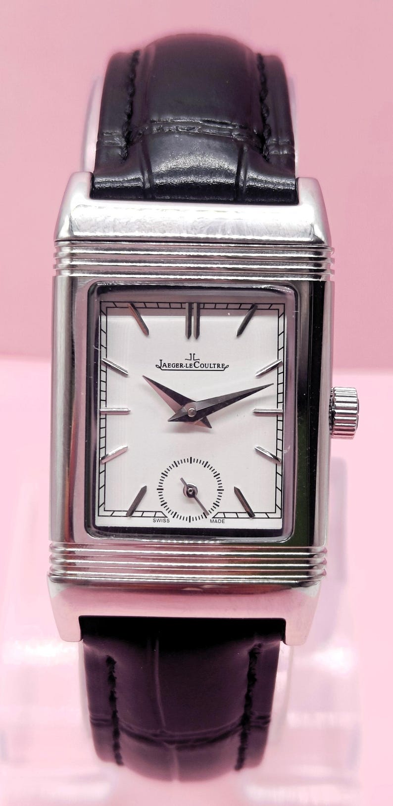 Pu&ograve; includere: Un orologio Jaeger-LeCoultre rettangolare argentato con quadrante bianco e lancette nere. L'orologio presenta un piccolo quadrante dei secondi e un cinturino in pelle nera. Il quadrante mostra il logo Jaeger-LeCoultre.