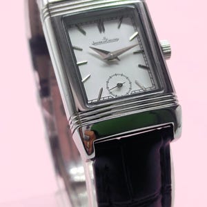 Pu&ograve; includere: Un orologio da polso rettangolare argentato con quadrante bianco e cinturino in pelle nera. Il quadrante dell'orologio presenta il marchio "JAEGER-LECOULTRE" e un piccolo quadrante dei secondi. L'orologio ha lancette e indici argentati.