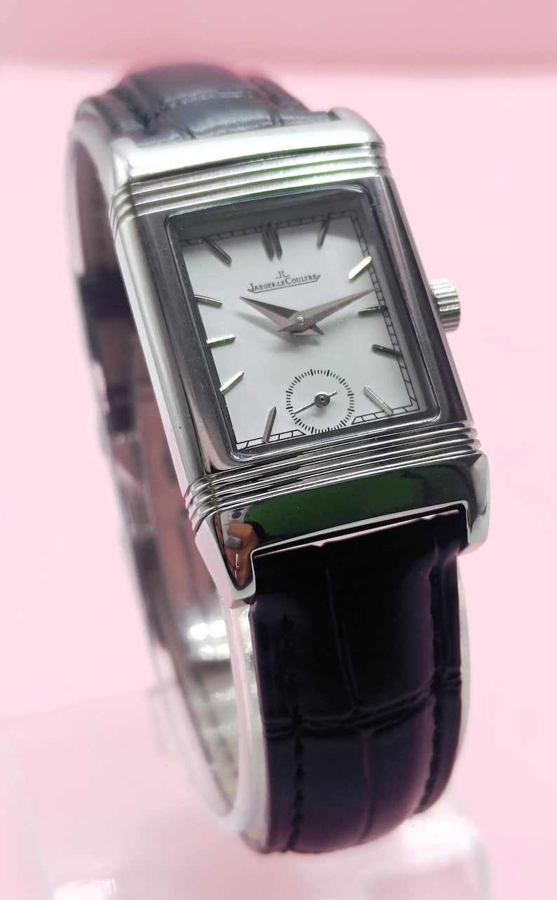 Pu&ograve; includere: Un orologio da polso rettangolare argentato con quadrante bianco e cinturino in pelle nera. Il quadrante dell'orologio presenta il marchio "JAEGER-LECOULTRE" e un piccolo quadrante dei secondi. L'orologio ha lancette e indici argentati.
