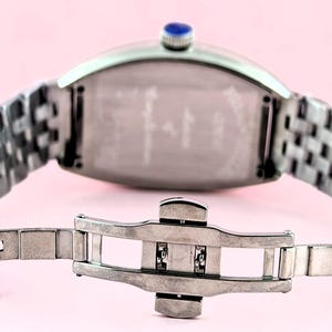 May include: Silver-toned wristwatch with a rectangular face and a woven metal band. The watch features a blue gem on the crown and a clasp closure. The watch is displayed against a pink background.