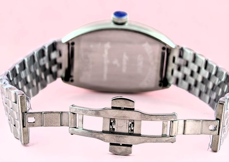 May include: Silver-toned wristwatch with a rectangular face and a woven metal band. The watch features a blue gem on the crown and a clasp closure. The watch is displayed against a pink background.