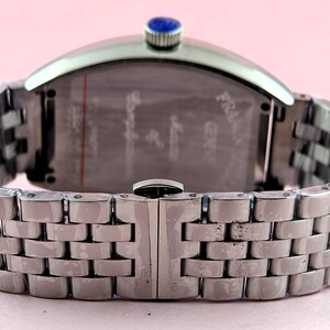 May include: Silver-toned wristwatch with a rectangular face and a linked metal band. The watch has a blue stone on the crown. The back of the watch has etched text. The band is closed with a clasp.