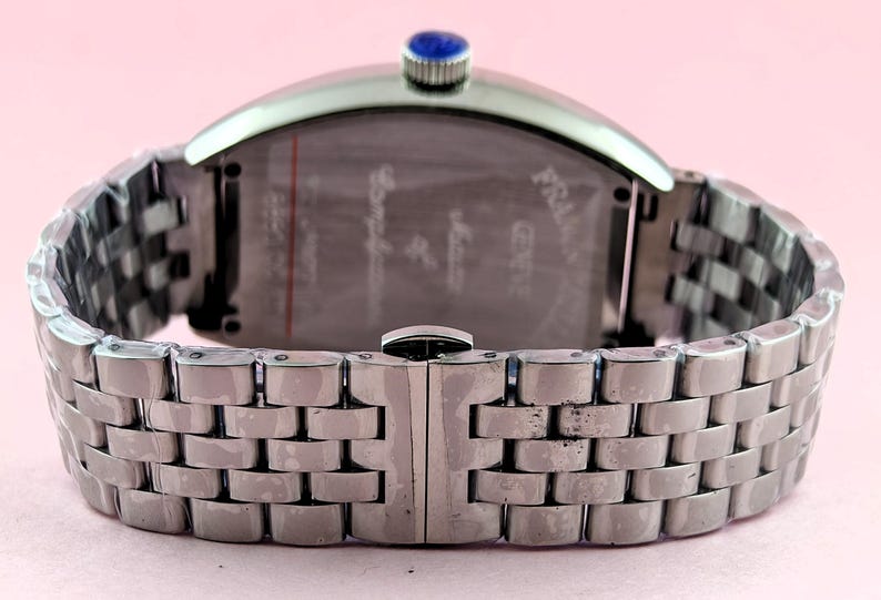 May include: Silver-toned wristwatch with a rectangular face and a linked metal band. The watch has a blue stone on the crown. The back of the watch has etched text. The band is closed with a clasp.