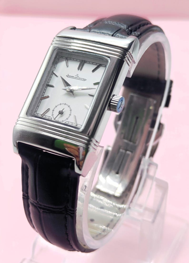 Pu&ograve; includere: Un orologio da polso rettangolare argentato con quadrante bianco e cinturino in pelle nera. L'orologio presenta un piccolo quadrante dei secondi e il marchio "Jaeger-LeCoultre". Il cinturino &egrave; nero e l'orologio &egrave; esposto su un supporto trasparente.
