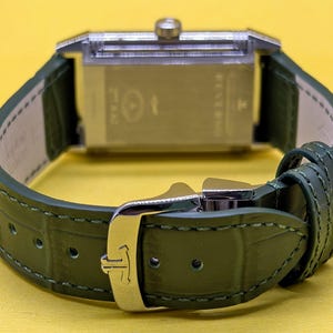 May include: A rectangular silver-toned watch case with a crown at the top, attached to a dark green textured leather strap. The strap features a silver-toned metal buckle with a logo and matching green stitching along the edges of the band.
