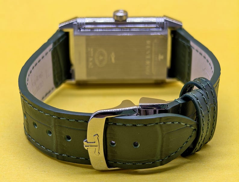May include: A rectangular silver-toned watch case with a crown at the top, attached to a dark green textured leather strap. The strap features a silver-toned metal buckle with a logo and matching green stitching along the edges of the band.