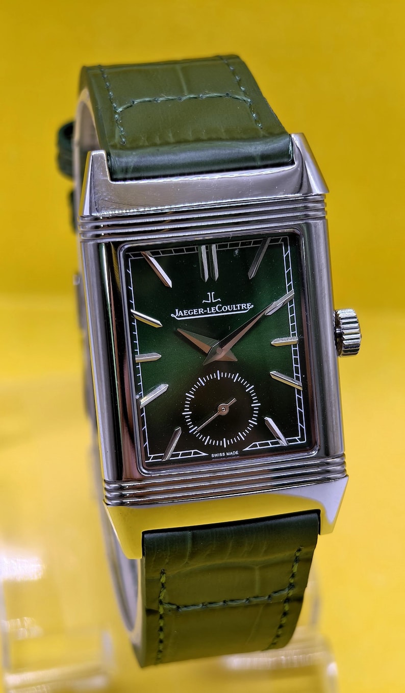 May include: A rectangular silver-tone wristwatch featuring a green gradient dial with silver hour markers, a small seconds sub-dial, and the text Jaeger-LeCoultre and Swiss Made. The watch has a green leather strap with a textured crocodile pattern.