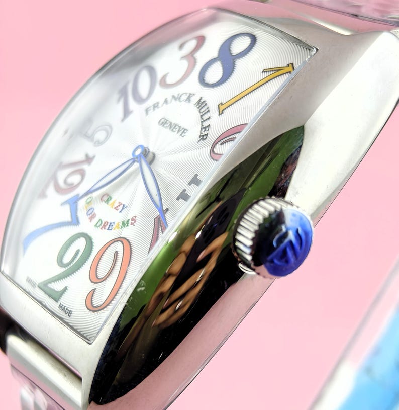 May include: A silver wristwatch with a white face featuring colorful numerals and hands. The watch face displays the text "FRANCK MULLER GENEVE" and "CRAZY COLOR DREAMS". The watch has a blue crown.