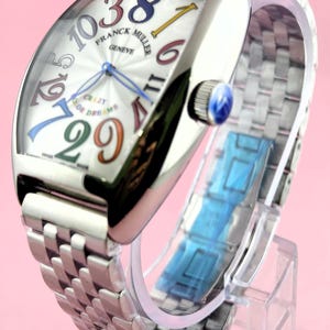 May include: A silver Franck Muller Geneve watch with a curved rectangular face. The watch face features large, colorful numerals and the words "CRAZY OR DREAMS". The watch has a silver band and a blue crown.