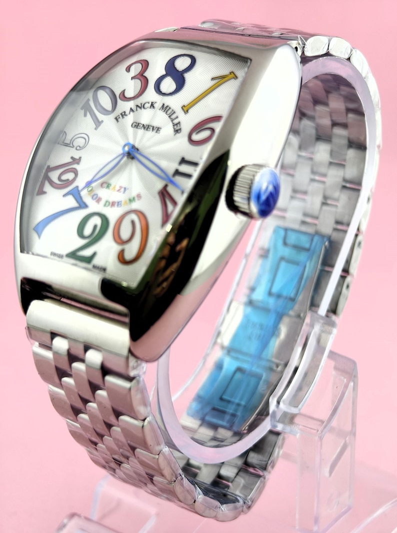 May include: A silver Franck Muller Geneve watch with a curved rectangular face. The watch face features large, colorful numerals and the words "CRAZY OR DREAMS". The watch has a silver band and a blue crown.