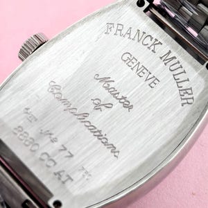May include: Close-up of the back of a silver-toned Franck Muller watch. The watch case is rectangular with rounded edges. The text "FRANCK MULLER GENEVE Master of Complications" is engraved on the back. The watch band is visible at the top.
