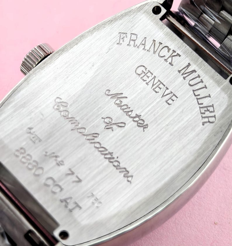 May include: Close-up of the back of a silver-toned Franck Muller watch. The watch case is rectangular with rounded edges. The text "FRANCK MULLER GENEVE Master of Complications" is engraved on the back. The watch band is visible at the top.