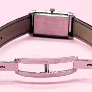 Pu&ograve; includere: Una chiusura per orologio argentata con l'iscrizione "JAEGER-LECOULTRE SWISS MADE" &egrave; mostrata. Il cinturino dell'orologio &egrave; in pelle nera con interno marrone chiaro. Il quadrante dell'orologio &egrave; rettangolare e argentato. Lo sfondo &egrave; rosa chiaro.