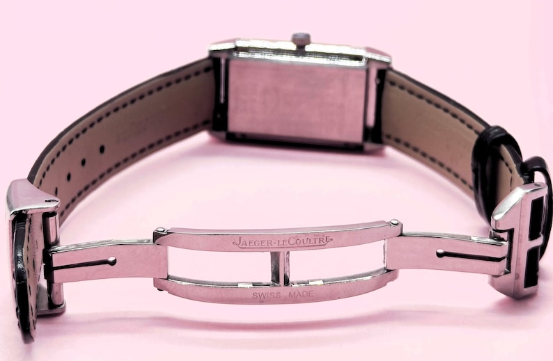 Pu&ograve; includere: Una chiusura per orologio argentata con l'iscrizione "JAEGER-LECOULTRE SWISS MADE" &egrave; mostrata. Il cinturino dell'orologio &egrave; in pelle nera con interno marrone chiaro. Il quadrante dell'orologio &egrave; rettangolare e argentato. Lo sfondo &egrave; rosa chiaro.