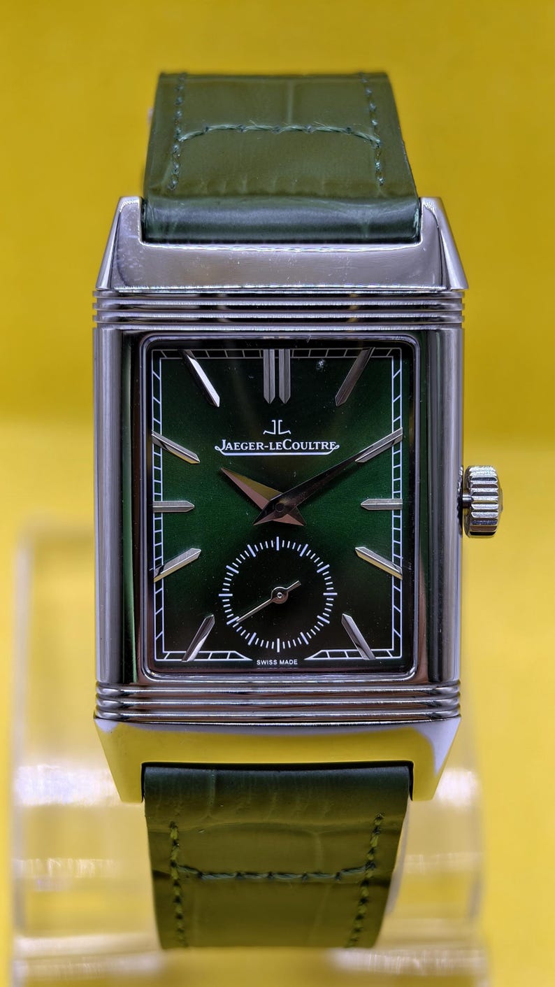 May include: A rectangular silver-tone watch with a green dial and a green leather strap. The dial features the text JAEGER-LECOULTRE and SWISS MADE, with silver-tone hour markers, a small seconds sub-dial, and a crown on the right side of the case.