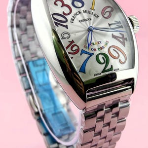 May include: A silver-toned wristwatch with a unique face featuring large, colorful numerals. The watch face displays the brand name "FRANCK MULLER GENEVE" and the words "CRAZY COLOR DREAMS". The watch has a silver band and a crown.