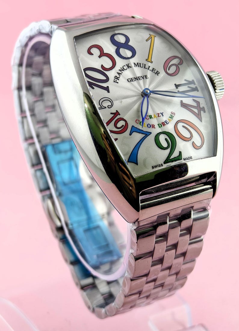 May include: A silver-toned wristwatch with a unique face featuring large, colorful numerals. The watch face displays the brand name "FRANCK MULLER GENEVE" and the words "CRAZY COLOR DREAMS". The watch has a silver band and a crown.