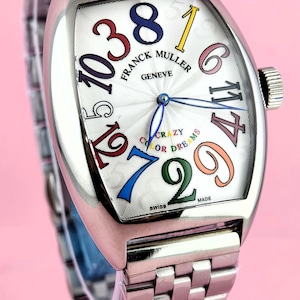 May include: A stainless steel Franck Muller Geneve watch with a white face and colorful numerals. The watch features a silver band and a unique, curved rectangular shape. The face includes the words "Crazy Color Dreams".