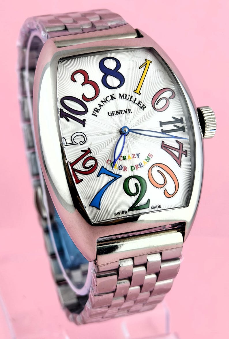 May include: A stainless steel Franck Muller Geneve watch with a white face and colorful numerals. The watch features a silver band and a unique, curved rectangular shape. The face includes the words "Crazy Color Dreams".