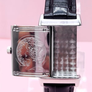 Pu&ograve; includere: Un orologio da polso rettangolare argentato con cinturino in pelle nera. Il quadrante dell'orologio presenta un disegno inciso del profilo di una donna. L'orologio &egrave; posto su uno sfondo rosa.