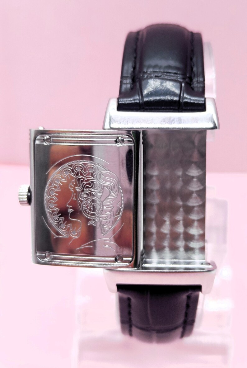 Pu&ograve; includere: Un orologio da polso rettangolare argentato con cinturino in pelle nera. Il quadrante dell'orologio presenta un disegno inciso del profilo di una donna. L'orologio &egrave; posto su uno sfondo rosa.
