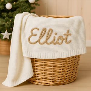 May include: A cream-colored knitted blanket with the name "Elliot" embroidered in a light brown rope-like font. The blanket is draped over a woven wicker basket. A Christmas tree is visible in the background.