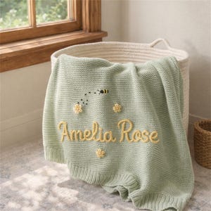 Può includere: Una coperta lavorata a maglia color verde salvia con il nome Amelia Rose ricamato in corsivo giallo, decorata con tre piccoli fiori gialli e un'ape nera e gialla con una scia di volo tratteggiata ricamata sopra il nome.