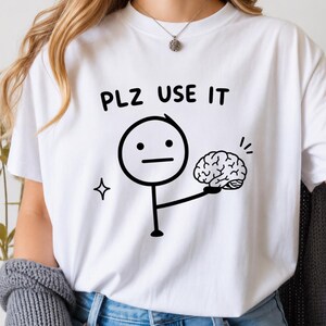 Puede incluir: Una camiseta blanca con un gráfico negro de un monigote sosteniendo un cerebro. Sobre la figura, el texto dice PLZ USE IT en una fuente negra sencilla. Un pequeño icono de estrella de cuatro puntas se sitúa a la izquierda del monigote.