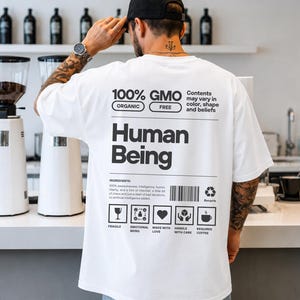 Könnte beinhalten: Ein weißes kurzärmeliges T-Shirt mit einem schwarzen Grafikdruck auf der Rückseite. Das Design imitiert ein Nährwertetikett mit dem Text "100% GMO FREE", "Human Being", einer Zutatenliste sowie Symbolen für zerbrechlich, emotional und Kaffee.