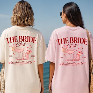 Könnte beinhalten: Zwei kurzärmelige T-Shirts, eines in Creme und eines in Hellrosa, mit einem roten Aufdruck auf der Rückseite. Das Design zeigt den Text THE BRIDE Club, EST. 2026 und Bachelorette party sowie Illustrationen von zwei Cocktails mit Zitrusfrüchten.