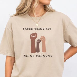 以下が含まれることがあります： ベージュの半袖Tシャツ。前面には黒い文字で「FASCHISMUS IST」と書かれ、その下に様々な肌の色の3つの拳を突き上げたイラストがあり、さらにその下に黒い文字で「KEINE MEINUNG」とプリントされたグラフィックデザインが施されています。