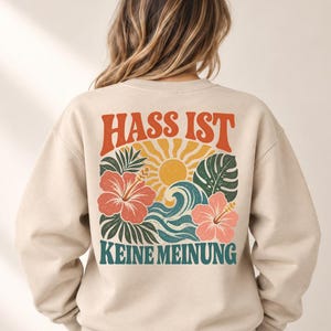 Op de afbeelding: Een beige sweater met ronde hals en een rugprint met de tekst HASS IST KEINE MEINUNG in oranje en blauwgroen. Het ontwerp bevat een gele zon, blauwe oceaangolven, groene tropische bladeren en roze hibiscusbloemen op de achterkant.