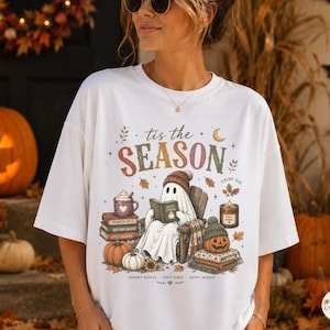 May include: A white t-shirt featuring a graphic of a ghost reading a book in a chair, surrounded by pumpkins, a mug, and a candle. The text reads 'tis the SEASON' with smaller text below including 'SPOOKY NIGHTS', 'COZY VIBES', and 'HAPPY HEARTS'.