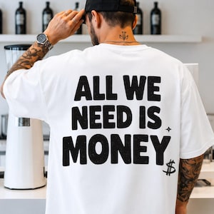 以下が含まれることがあります： 背面にALL WE NEED IS MONEYという太字の黒いテキストがプリントされた白い半袖Tシャツ。グラフィックの最後の単語の末尾には、小さな4つの光芒を持つ星のアイコンとドル記号が添えられています。